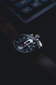 A close-up view of a luxury wristwatch with a dark leather band and a detailed dial. The watch features metallic elements and a prominent winding crown. In the background, a keyboard with highlighted keys is visible, blending with the dimly lit setting.