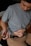 A person wearing a gray t-shirt and brown pants is sitting and holding an injection pen. They appear to be administering an injection in the abdomen while holding the skin.
