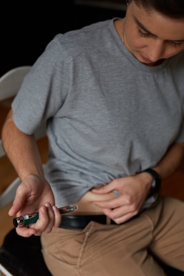 A person wearing a gray t-shirt and brown pants is sitting and holding an injection pen. They appear to be administering an injection in the abdomen while holding the skin.