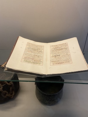 An open book with handwritten Arabic script on both visible pages, positioned in a display case. The book is placed on a glass shelf, surrounded by some artifacts including a metal chain and a cylindrical object beneath it.