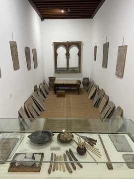 A narrow room in a museum or historical site contains various historical artifacts arranged along the walls and in a central display case. The walls are adorned with several rectangular stone slabs featuring inscriptions or carvings. These slabs are also neatly lined along both sides of the floor. A small table covered with a cloth is positioned at the far end of the room under a decorative wooden arch or window frame. In the foreground, within a glass display case, are several objects such as carved sticks, bowls, and tools, suggesting an exhibit of ancient scripts or writing implements.
