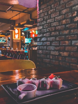 A cozy restaurant interior with dim lighting, featuring brick walls and wooden accents. In the foreground, a plate of sushi is elegantly presented with a small dish of soy sauce, placed on a dark wooden table. The background shows various colorful signs and ambient lights, creating a warm and inviting atmosphere.