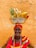 An elderly person wearing a bright, colorful outfit with a matching headscarf smiles warmly. They balance a silver bowl on their head, filled with tropical fruits including bananas, a pineapple, and a papaya. The background features a textured, ochre-colored wall.