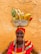 An elderly person wearing a bright, colorful outfit with a matching headscarf smiles warmly. They balance a silver bowl on their head, filled with tropical fruits including bananas, a pineapple, and a papaya. The background features a textured, ochre-colored wall.