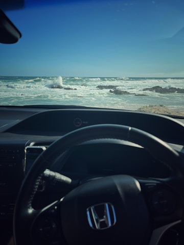 Close-up of hands gently holding a steering wheel with ocean waves in the background.