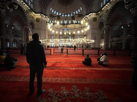 A spacious interior with ornate architectural details, featuring large chandeliers hanging from the ceiling, intricate arches, and vibrant red carpeting covering the floor. People are sitting and standing in various areas, likely engaged in prayer or contemplation.