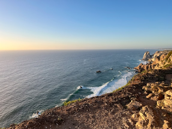 A coastal cliffside view capturing the expansive ocean meeting the rocky shore. The waves gently lap against the edge while rugged cliffs extend into the distance under a clear sky. The sun casts a warm glow, enhancing the natural beauty of the scene.