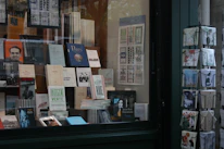 A bookstore window display with a variety of books and postcards. The books are neatly arranged on shelves with numerous titles visible, including travel guides and literature. Next to the display, there is a rotating stand filled with postcards featuring artistic and scenic designs.