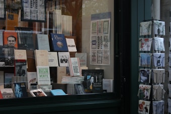 A bookstore window display with a variety of books and postcards. The books are neatly arranged on shelves with numerous titles visible, including travel guides and literature. Next to the display, there is a rotating stand filled with postcards featuring artistic and scenic designs.