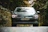 A car with sleek headlights and a prominent Volkswagen logo is parked on a road surrounded by lush green trees. The scene has a serene and secluded feeling, with the vehicle's modern design contrasting with the natural backdrop.