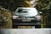 A car with sleek headlights and a prominent Volkswagen logo is parked on a road surrounded by lush green trees. The scene has a serene and secluded feeling, with the vehicle's modern design contrasting with the natural backdrop.