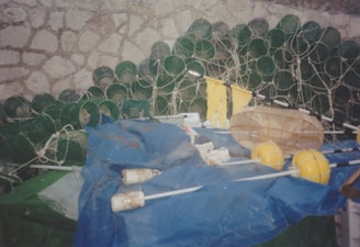 A collection of green cylindrical objects, likely fishing traps or cages, are stacked against a stone wall. In the foreground, there are yellow buoys, nets, and white objects placed on a blue tarp.