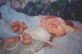 A collection of fishing equipment, including tangled nets with shades of orange and pink, is spread across a surface. Orange floats are attached to a long pole, and rolls of netting and other materials are scattered around. In the background, various cages or traps are visible against a stone or concrete wall.
