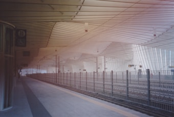 A modern train station platform with an architectural ceiling design featuring parallel lines and gentle curves. The platform is largely empty, with metal fencing separating the tracks. Various signs are visible, including those indicating accessibility features.