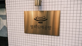 A brass sign with Korean text and a stylized design is mounted on a tiled wall. The wall features a grid pattern of small white tiles.