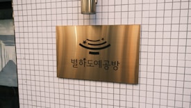 A brass sign with Korean text and a stylized design is mounted on a tiled wall. The wall features a grid pattern of small white tiles.