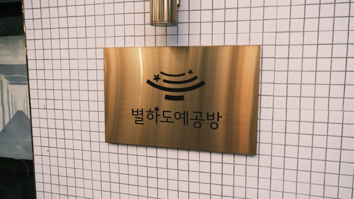 A brass sign with Korean text and a stylized design is mounted on a tiled wall. The wall features a grid pattern of small white tiles.