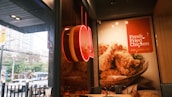 A fast-food restaurant with a large advertisement featuring a basket of fried chicken. The interior is decorated with a wooden panel and modern lighting, while a red neon sign is visible. The window shows a view of an urban street with cars and buildings outside.