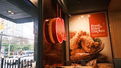 A fast-food restaurant with a large advertisement featuring a basket of fried chicken. The interior is decorated with a wooden panel and modern lighting, while a red neon sign is visible. The window shows a view of an urban street with cars and buildings outside.