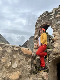 A person dressed in bright outdoor clothing, including an orange cap, yellow jacket, and red pants, is seen standing on an ancient stone structure with an arch. The stonework appears weathered and is surrounded by a mountainous landscape under a cloudy sky.