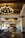 A luxurious hotel lobby featuring large, ornate chandeliers hanging from a wooden beam ceiling. The space is adorned with stylish furniture, including cushioned chairs and potted plants. The glossy tile floor reflects the warm lighting creating an inviting atmosphere.