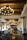 A luxurious hotel lobby featuring large, ornate chandeliers hanging from a wooden beam ceiling. The space is adorned with stylish furniture, including cushioned chairs and potted plants. The glossy tile floor reflects the warm lighting creating an inviting atmosphere.