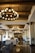 A luxurious hotel lobby featuring large, ornate chandeliers hanging from a wooden beam ceiling. The space is adorned with stylish furniture, including cushioned chairs and potted plants. The glossy tile floor reflects the warm lighting creating an inviting atmosphere.