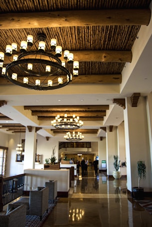 A luxurious hotel lobby featuring large, ornate chandeliers hanging from a wooden beam ceiling. The space is adorned with stylish furniture, including cushioned chairs and potted plants. The glossy tile floor reflects the warm lighting creating an inviting atmosphere.