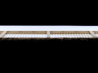 A geometric composition featuring an architectural structure with repetitive vertical slats and beams against a stark contrast of black and white tones. The upper portion of the image features a solid black background, while the structure itself is illuminated, highlighting the linear design.