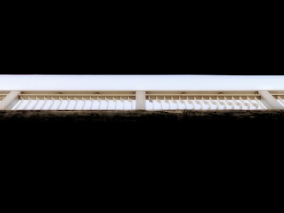 A geometric composition featuring an architectural structure with repetitive vertical slats and beams against a stark contrast of black and white tones. The upper portion of the image features a solid black background, while the structure itself is illuminated, highlighting the linear design.