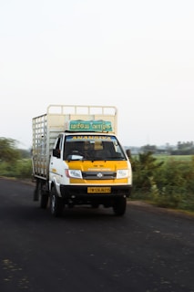 A yellow and white truck is driving down a rural road. The truck has a signboard with text in a script, and the vehicle appears to be moving at a moderate speed. There is greenery on both sides of the road, and the sky is clear.