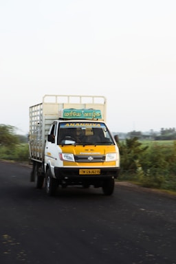 A yellow and white truck is driving down a rural road. The truck has a signboard with text in a script, and the vehicle appears to be moving at a moderate speed. There is greenery on both sides of the road, and the sky is clear.