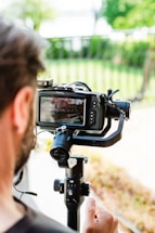 A person is holding a professional camera mounted on a gimbal stabilizer. The camera display shows the scene being captured. A blurred background with greenery and a railing can be seen.