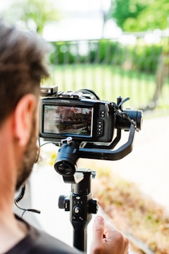 A person is holding a professional camera mounted on a gimbal stabilizer. The camera display shows the scene being captured. A blurred background with greenery and a railing can be seen.