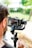 A person is holding a professional camera mounted on a gimbal stabilizer. The camera display shows the scene being captured. A blurred background with greenery and a railing can be seen.