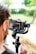 A person is holding a professional camera mounted on a gimbal stabilizer. The camera display shows the scene being captured. A blurred background with greenery and a railing can be seen.