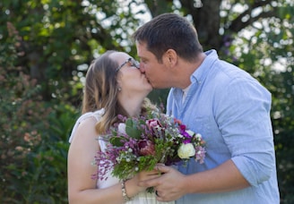 A couple engaged in a kiss, with the woman holding a vibrant bouquet of flowers. The background features lush green foliage, suggesting an outdoor setting. The man is wearing a light blue shirt, and the woman has long hair with glasses, dressed in white.