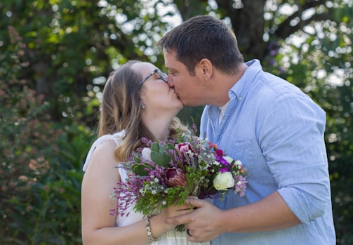 A couple engaged in a kiss, with the woman holding a vibrant bouquet of flowers. The background features lush green foliage, suggesting an outdoor setting. The man is wearing a light blue shirt, and the woman has long hair with glasses, dressed in white.