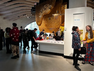 A dimly lit room filled with people engaging in activities around a large display. The display features a honeycomb structure on the wall, creating an impression of a beehive. Several individuals are gathered around a central table, reading and interacting with the exhibit. The room has a modern architecture with curved wooden beams on the ceiling, adding to the artistic ambiance.