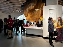 A dimly lit room filled with people engaging in activities around a large display. The display features a honeycomb structure on the wall, creating an impression of a beehive. Several individuals are gathered around a central table, reading and interacting with the exhibit. The room has a modern architecture with curved wooden beams on the ceiling, adding to the artistic ambiance.