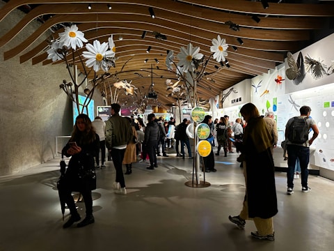 A busy exhibition space with a modern, wooden ceiling structure and large floral sculptures hanging overhead. People are walking around and observing various displays that are well-illuminated on the walls. Interactive panels and informational boards are present, contributing to an educational atmosphere.