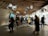 A busy exhibition space with a modern, wooden ceiling structure and large floral sculptures hanging overhead. People are walking around and observing various displays that are well-illuminated on the walls. Interactive panels and informational boards are present, contributing to an educational atmosphere.