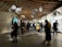 A busy exhibition space with a modern, wooden ceiling structure and large floral sculptures hanging overhead. People are walking around and observing various displays that are well-illuminated on the walls. Interactive panels and informational boards are present, contributing to an educational atmosphere.