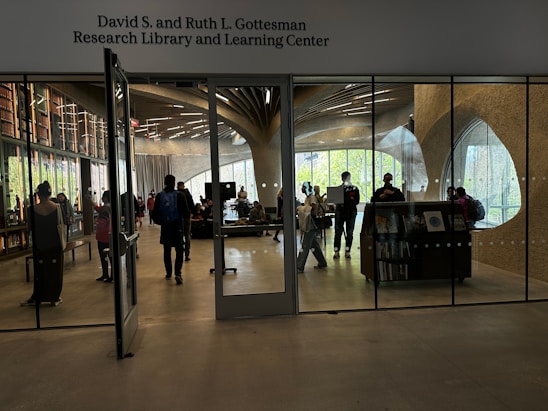A modern library and learning center with people interacting. The room is enclosed with large glass panels, showing bookshelves with numerous books. The walls have unique curved designs with large windows letting in natural light. Several individuals are walking or standing, some carrying backpacks.