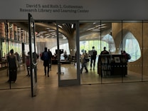 A modern library and learning center with people interacting. The room is enclosed with large glass panels, showing bookshelves with numerous books. The walls have unique curved designs with large windows letting in natural light. Several individuals are walking or standing, some carrying backpacks.