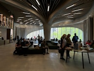 A modern interior space with curved, organic architectural features. People are seated and standing around tables, engaging in activities like reading or browsing. Large windows allow natural light to illuminate the room, and the ceiling features an elaborate, artistic design with wood-like elements and embedded lights.