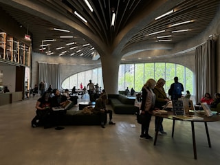 A modern interior space with curved, organic architectural features. People are seated and standing around tables, engaging in activities like reading or browsing. Large windows allow natural light to illuminate the room, and the ceiling features an elaborate, artistic design with wood-like elements and embedded lights.