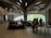 A modern interior space with curved, organic architectural features. People are seated and standing around tables, engaging in activities like reading or browsing. Large windows allow natural light to illuminate the room, and the ceiling features an elaborate, artistic design with wood-like elements and embedded lights.