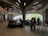 A modern interior space with curved, organic architectural features. People are seated and standing around tables, engaging in activities like reading or browsing. Large windows allow natural light to illuminate the room, and the ceiling features an elaborate, artistic design with wood-like elements and embedded lights.