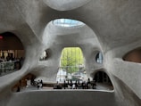 Futuristic architectural interior with organic, curving walls made of textured concrete. The space features large, irregular windows offering views of greenery outside. A glass ceiling allows natural light to illuminate the large open area, where groups of people are gathered, some looking out the windows and others congregating near an information desk.